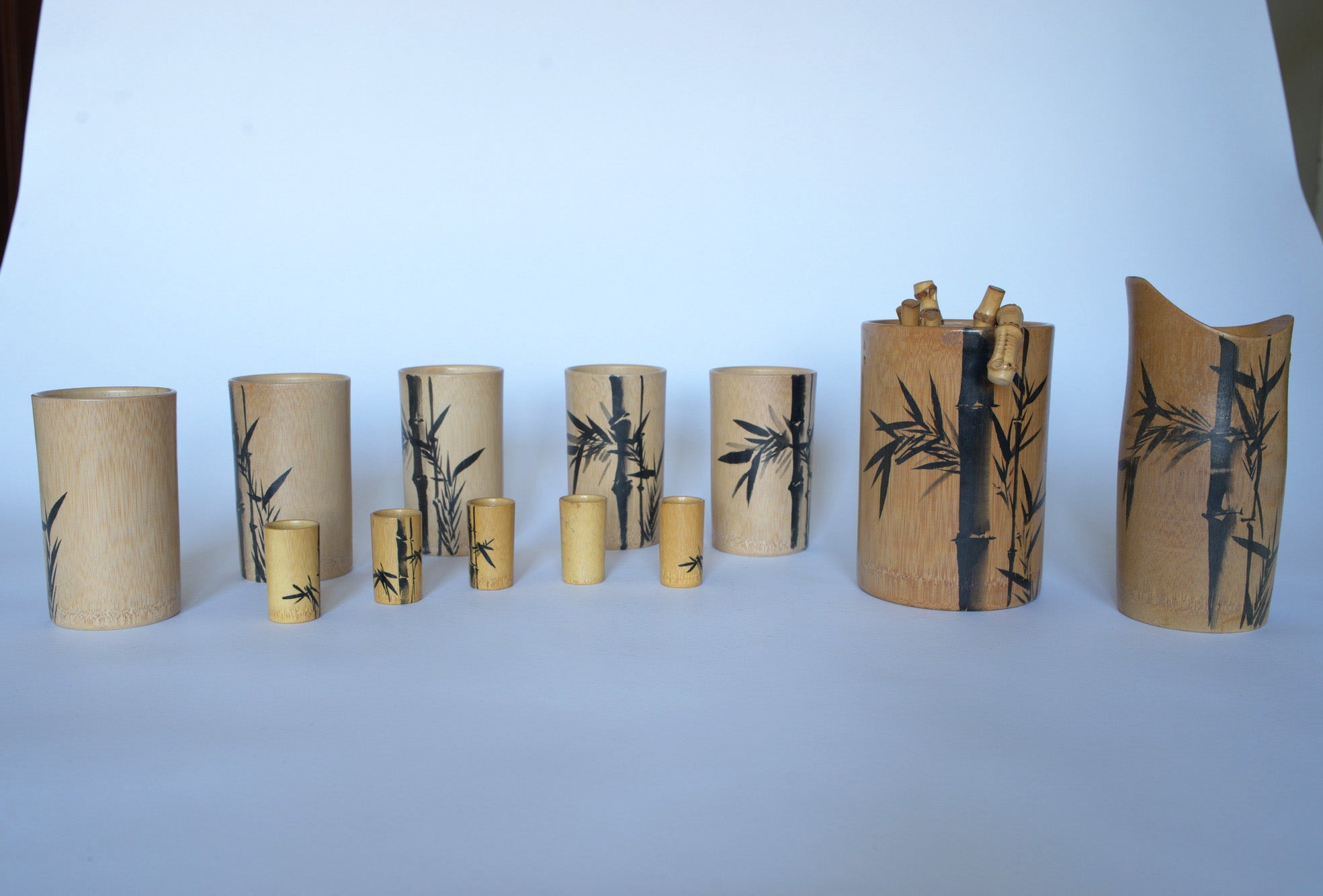 The Euro Trash Vintage Bamboo Motif Wooden Barware Set features wooden cups and containers with black bamboo designs, arranged in a row on a white surface. Ideal for mid-century tropical decor, some pieces hold short cylindrical items.