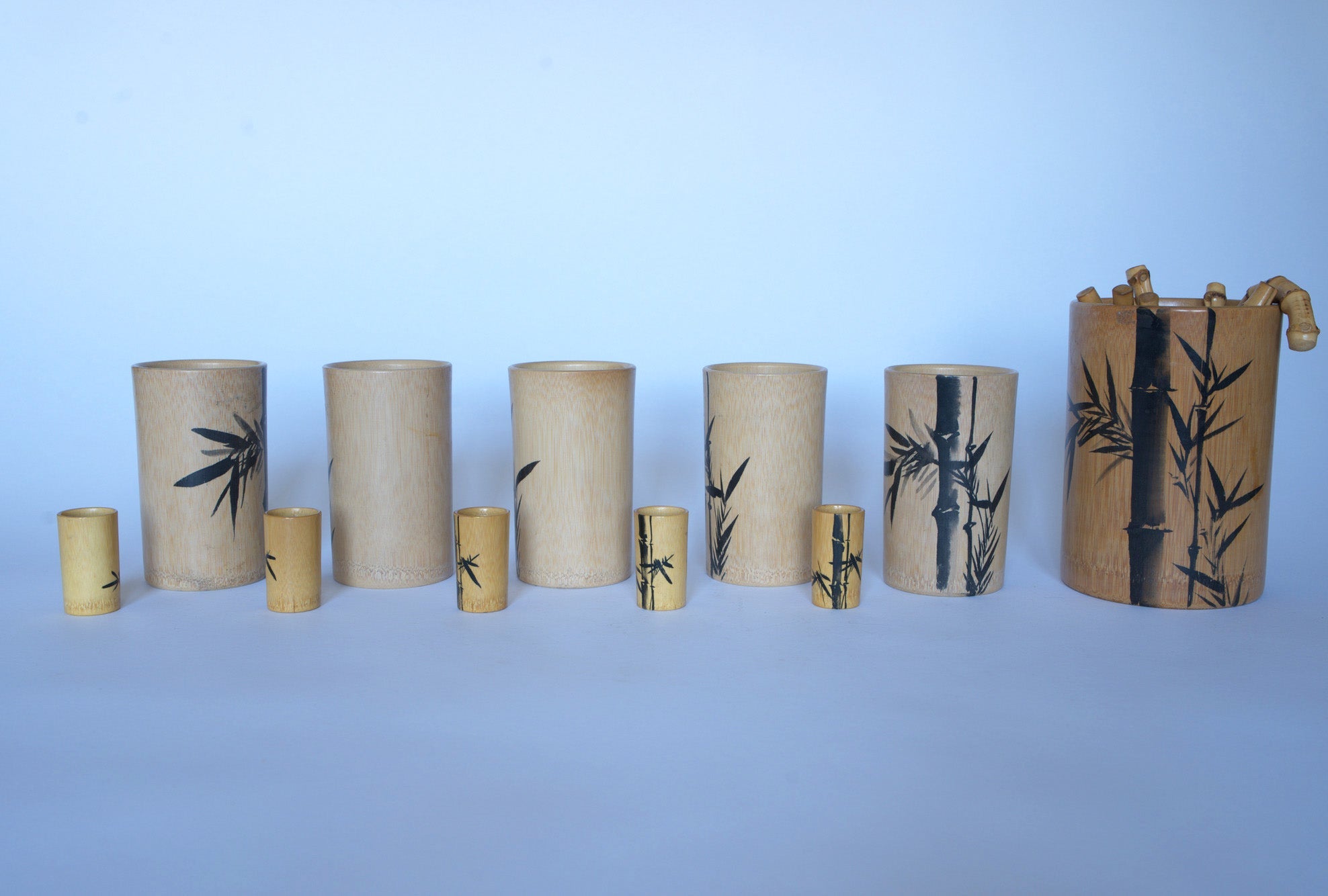 The Euro Trash Vintage Bamboo Motif Wooden Barware Set features cylindrical wooden cups in assorted sizes with painted bamboo leaf designs, displayed on a white background—ideal for adding mid-century tropical charm to your décor.