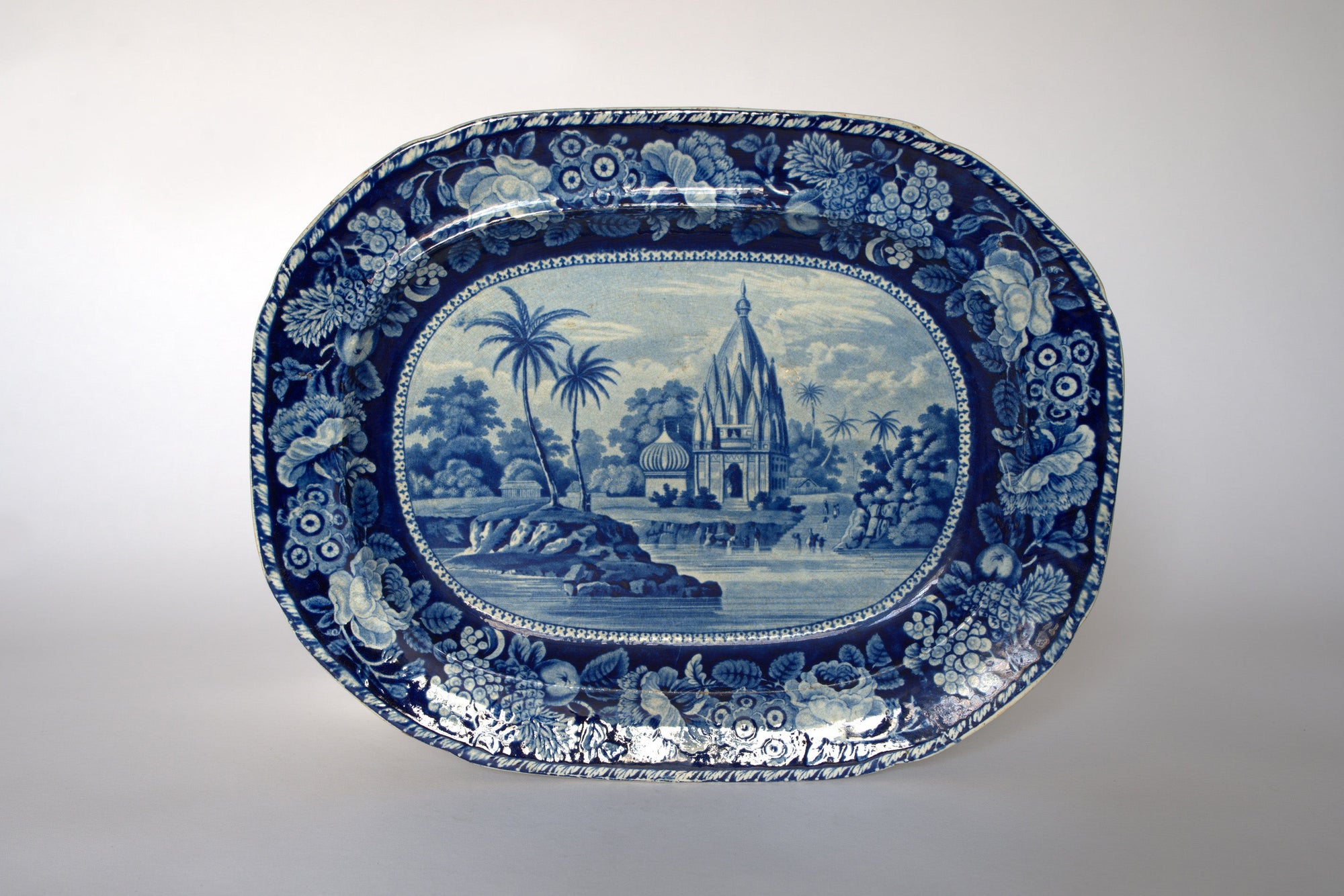 The Euro Trash Vintage Blue & White Floral Transferware Serving Platter features a central temple scene by a river with trees and people, framed by a floral border—ideal as a unique vintage serving piece or decorative accent.