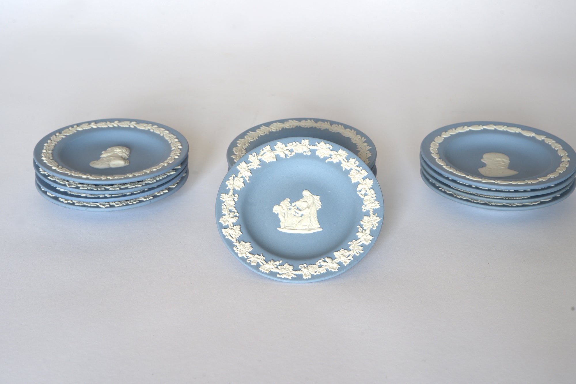 Euro Trash Vintage Blue Jasperware Style Relief Plates: six blue plates with white neoclassical designs—four stacked left, three right, plus one central decorative plate featuring a white relief of figures.