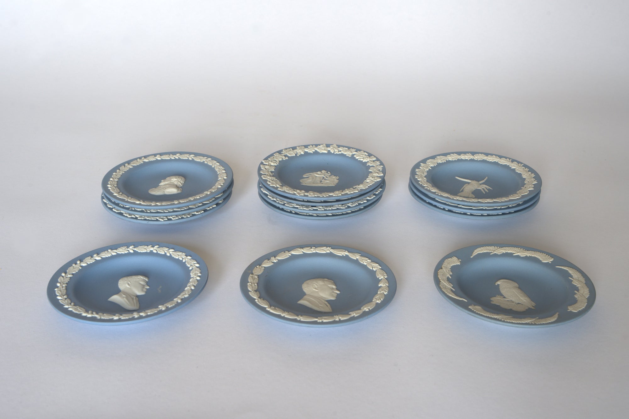 Seven Euro Trash Vintage Blue Jasperware Style Relief Plates, adorned with profiles, animals, and floral motifs, are arranged in three rows on a light background to form an elegant decorative wall display set.