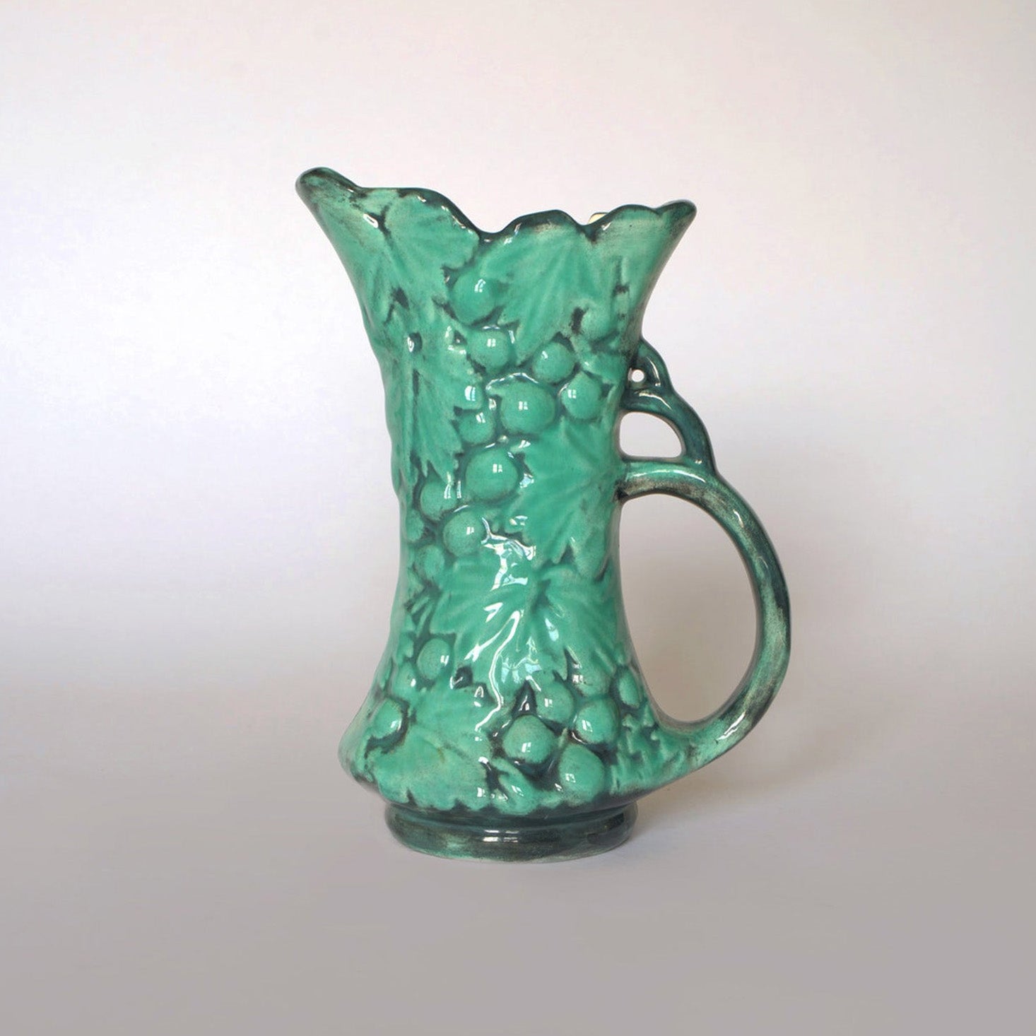 The Euro Trash Vintage Green Majolica Style Grape Motif Ceramic Pitcher features a glossy finish, raised grapevine design, scalloped rim, and curved handle for timeless vintage appeal.