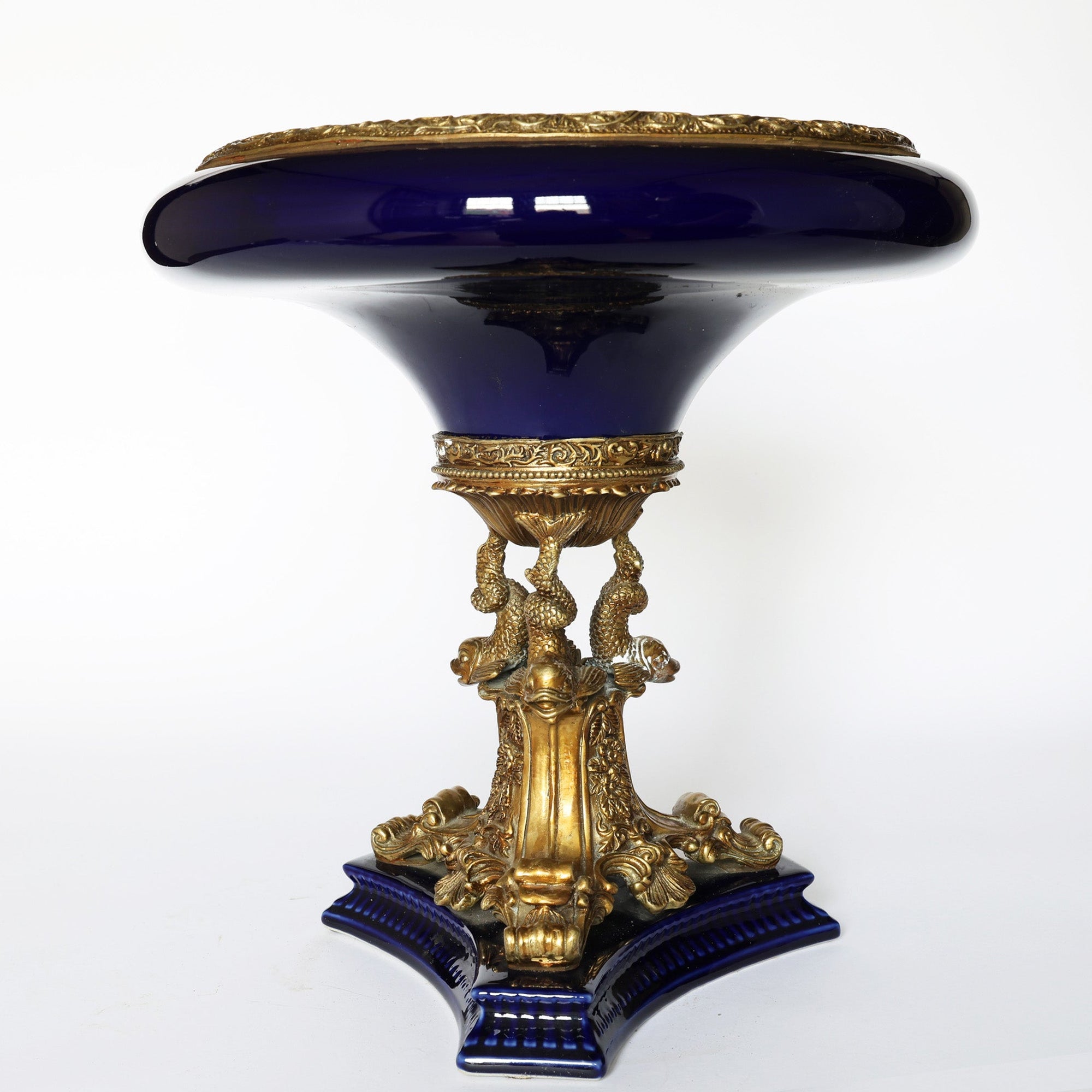 The Euro Trash Antique Cobalt Porcelain & Gilt Bronze Centerpiece Tazza features a glossy cobalt bowl top, ornate gilt bronze details, sculpted gold base with figures and patterns, and stands on a blue four-cornered platform.