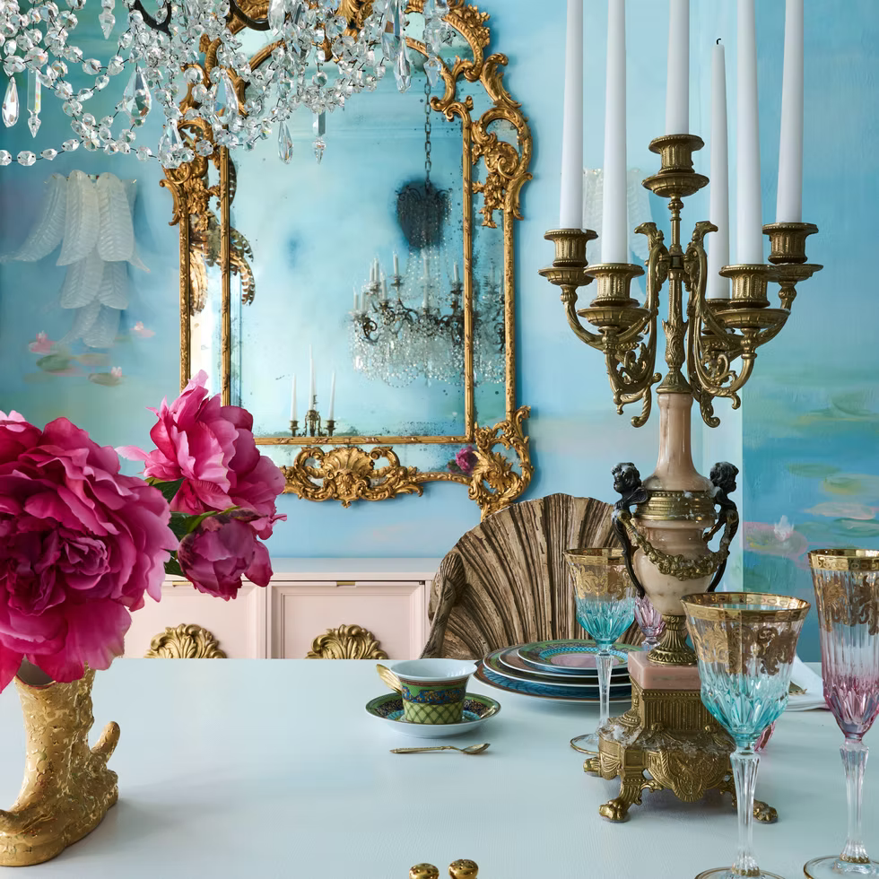 An ornate dining table set with gold-trimmed glassware, a large gold candelabra, pink flowers in a decorative vase, and an elaborate gilded mirror reflecting a crystal chandelier, all against a blue wall.