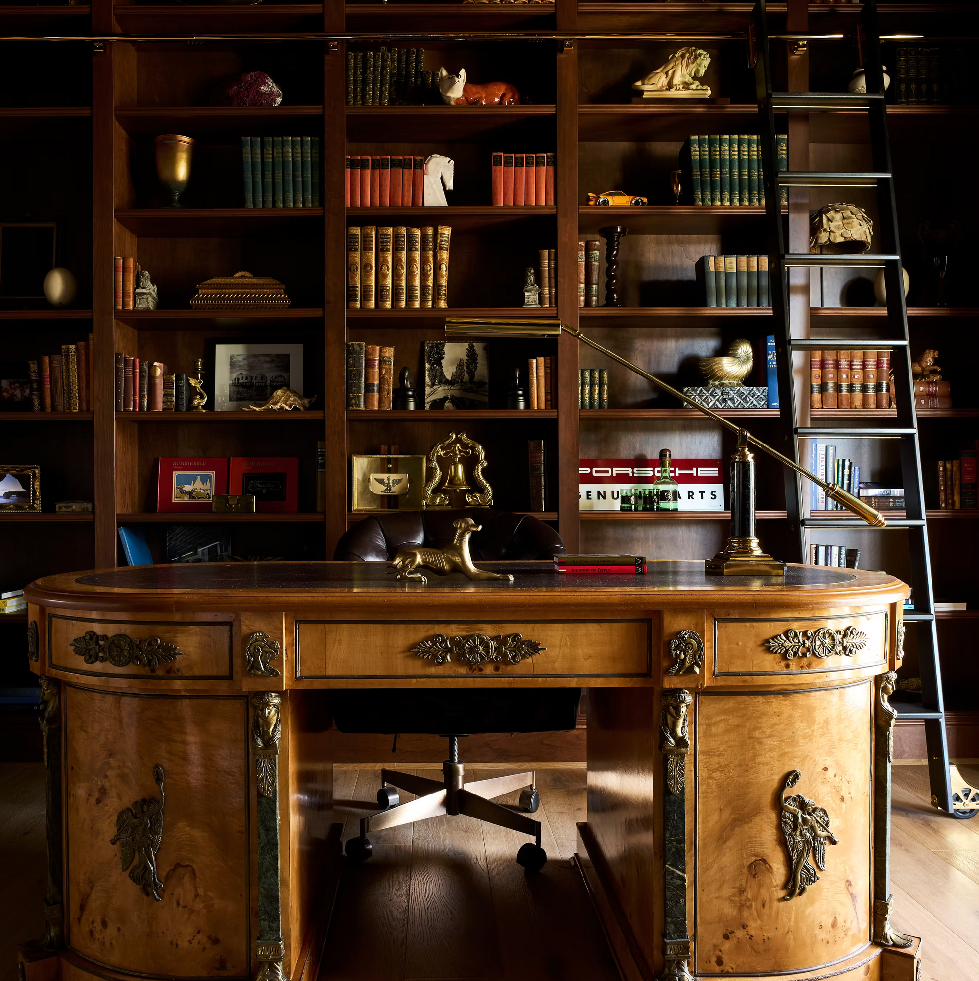 A luxurious wooden office with ornate desk, leather chair, and large bookshelf filled with books, art, and collectibles. A rolling ladder leans on the shelves; a gold lamp and decor items sit on the desk.