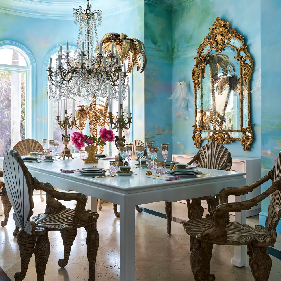 An elegant dining room with ocean-themed decor, featuring shell-shaped chairs, a white table set with pink flowers, a crystal chandelier, ornate gold-framed mirror, and blue walls with underwater murals.