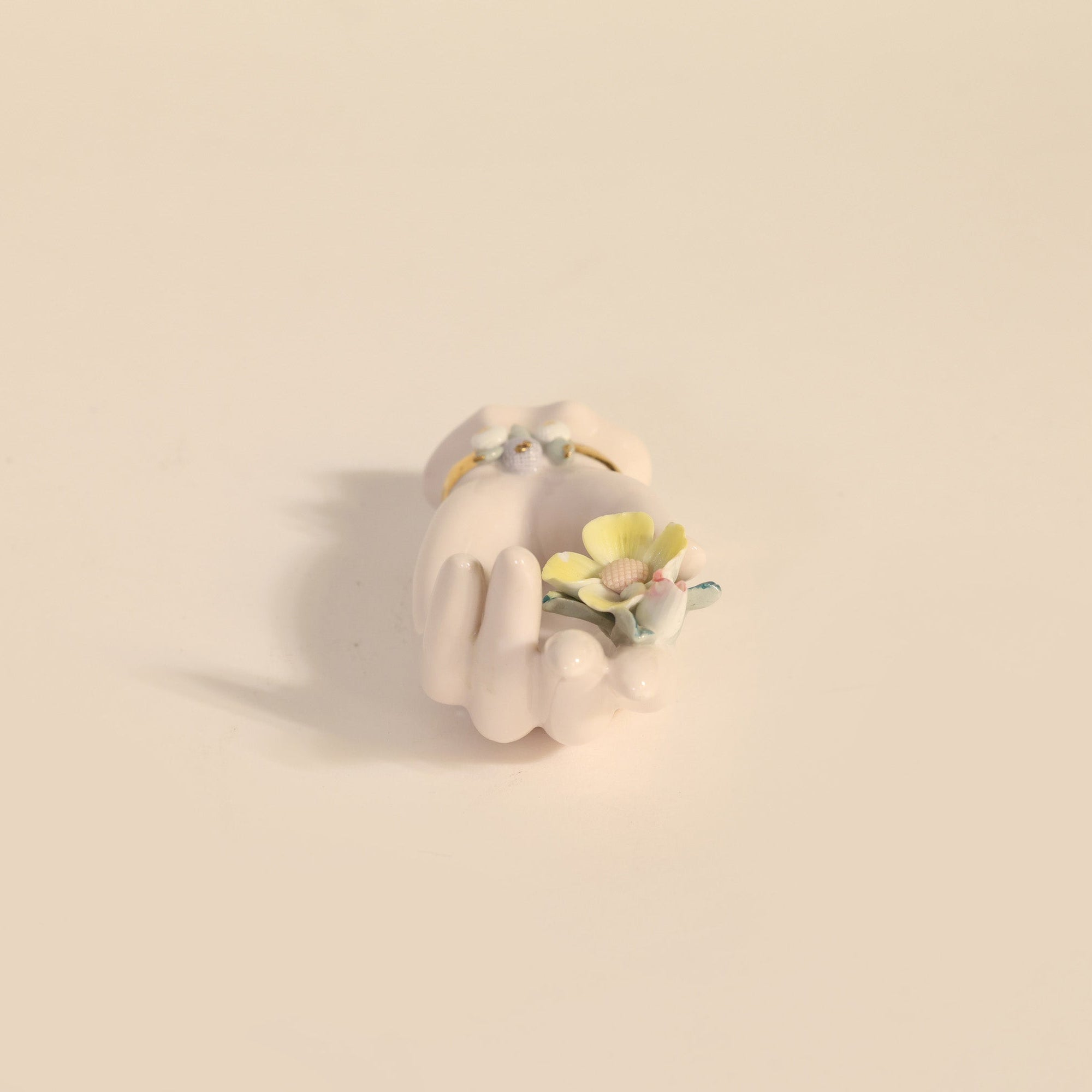 Small white ceramic hand figurine with yellow flowers on a beige background.