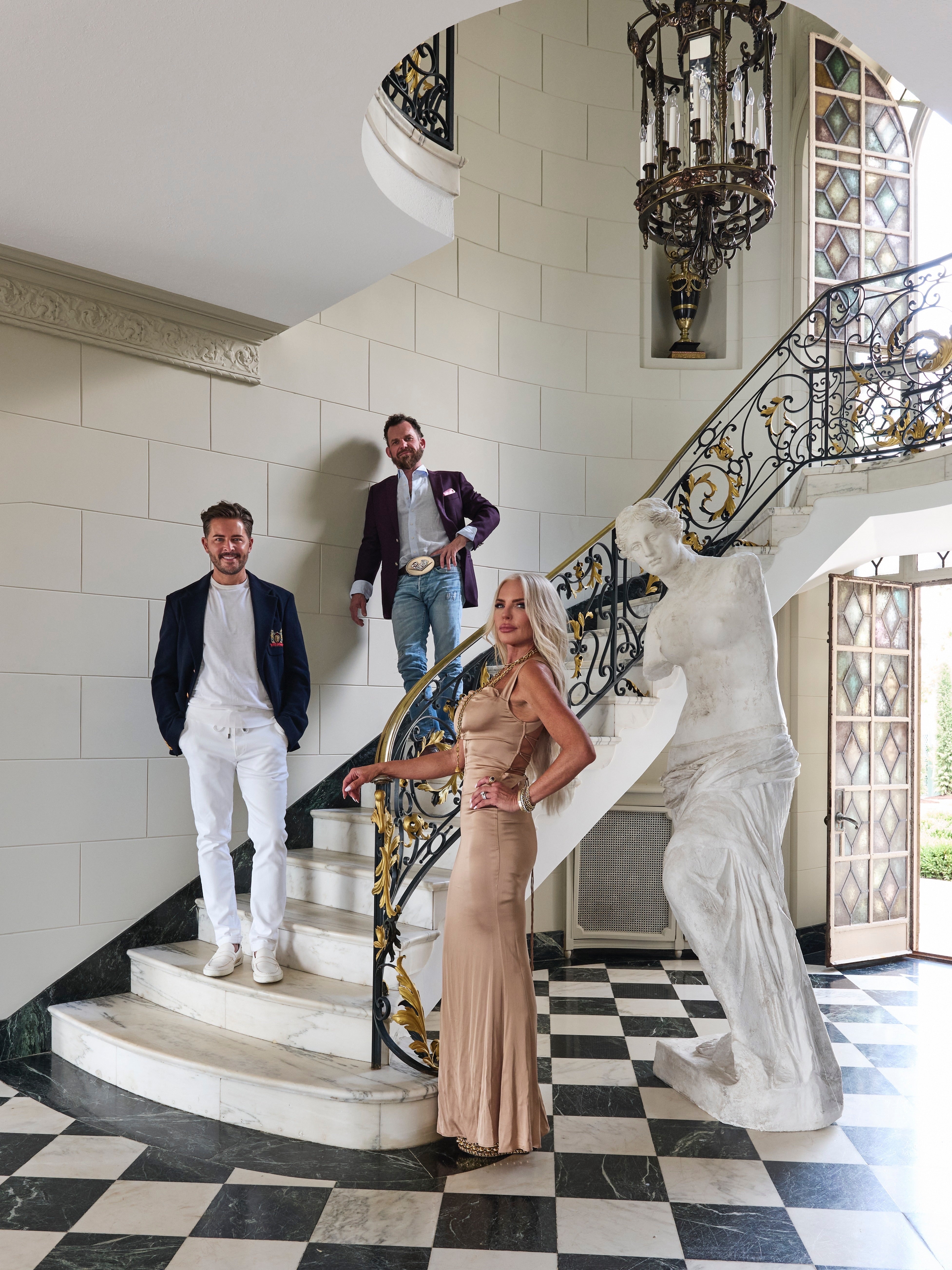 Three people pose on a marble staircase in an elegant foyer with checkered floors, ornate railings, a large statue, and tall windows. Two men stand on the steps and a woman in a gold dress stands in front.